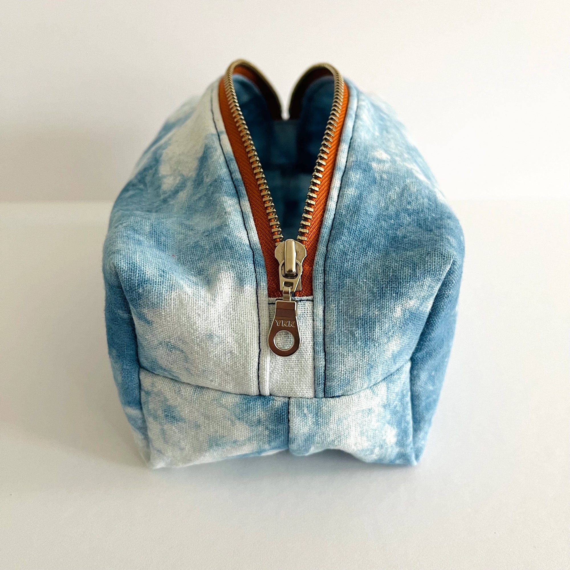 The Faire Dyed Toiletries Bag is a small, box-shaped pouch with natural blue and white tie-dye, brown zipper trim, and a silver pull, shown on a plain white background.