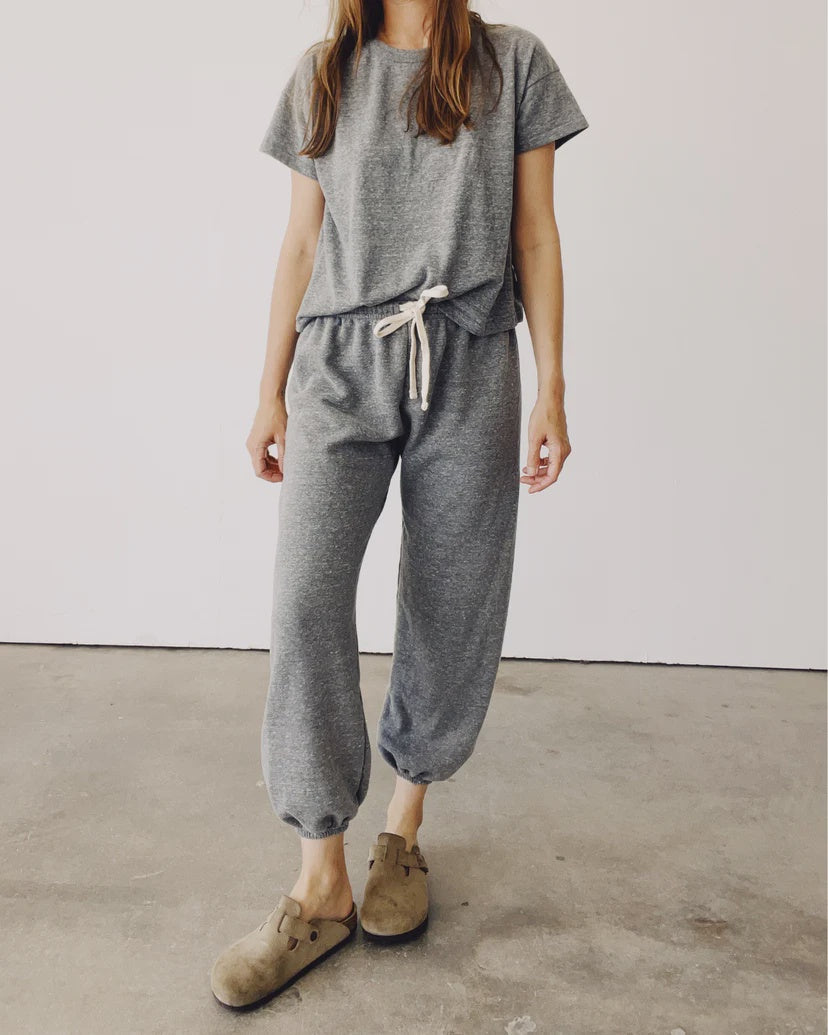 A person dressed in The Sweatpants by Hey Gang, featuring an elastic waistband and ankle cuffs, along with a matching gray French terry T-shirt, stands on a concrete floor against a plain white wall. They are wearing tan slip-on shoes with buckles, and the top is partially tucked in.