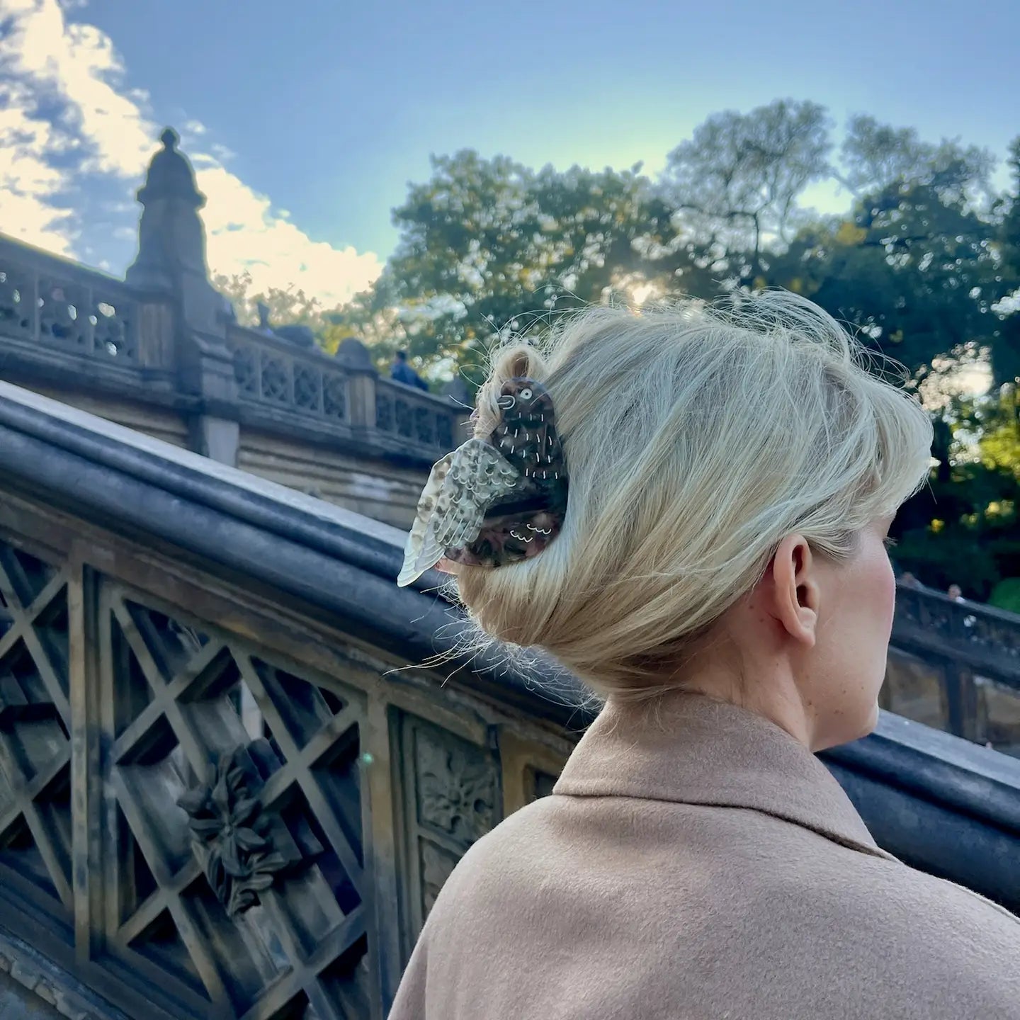A woman with blonde hair pinned up by Faire’s Hand Painted Pigeon Clip stands near an ornate stone staircase outdoors, framed by trees and a partly cloudy sky.