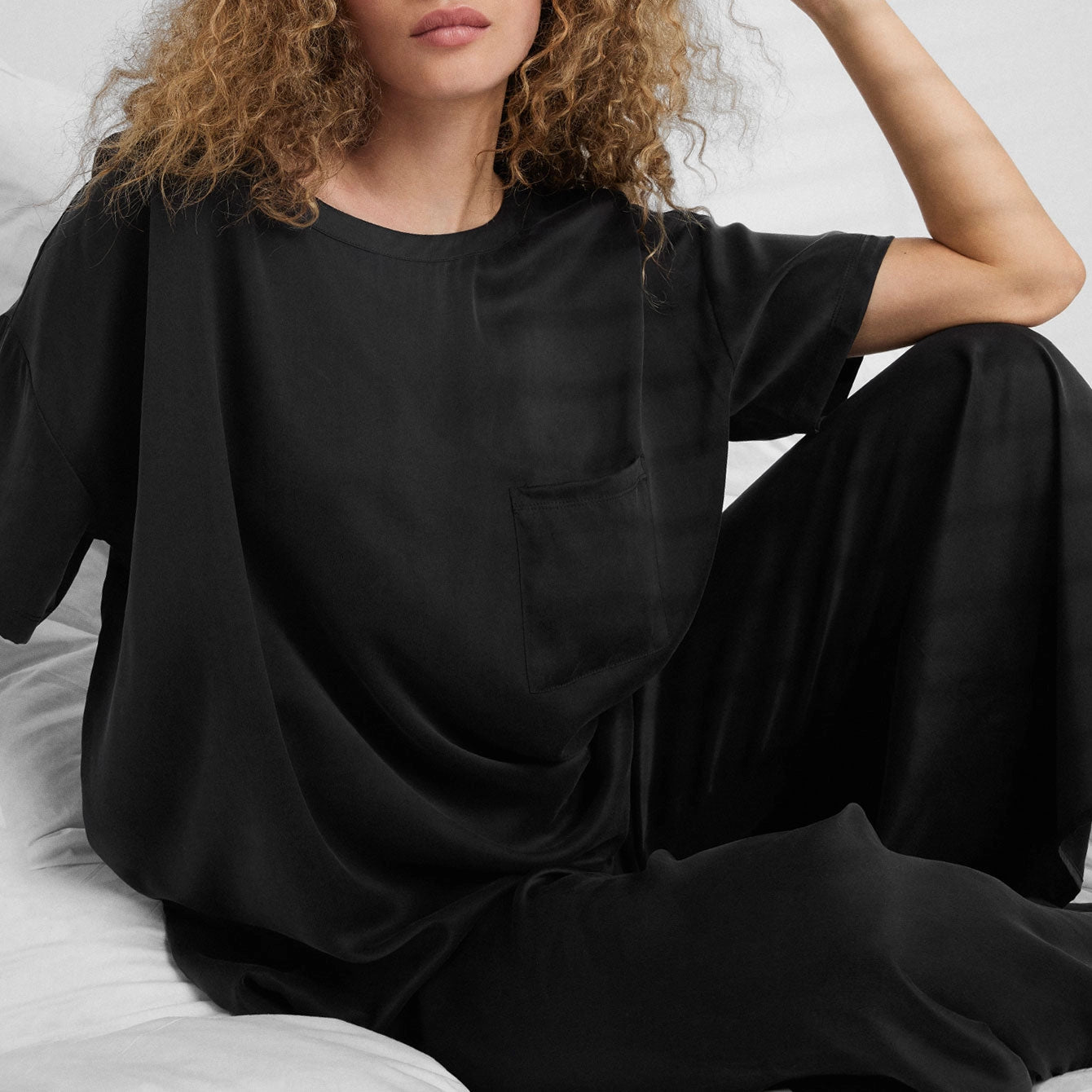 A woman with curly hair sits on a bed wearing the Faire Lunya Washable Silk Tee. Only her upper body and part of her face are visible, against a background of white bedding.