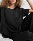 A woman with curly hair sits on a bed wearing the Faire Lunya Washable Silk Tee. Only her upper body and part of her face are visible, against a background of white bedding.