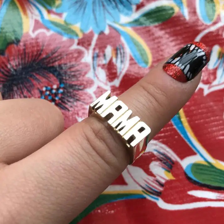 A hand wears Faire's Mama Ring, a brass ring spelling "MAMA" on the middle finger. The shiny ring pops against floral patterns, while the fingernail features black, red, and white nail art.