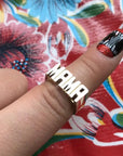 A hand wears Faire's Mama Ring, a brass ring spelling "MAMA" on the middle finger. The shiny ring pops against floral patterns, while the fingernail features black, red, and white nail art.