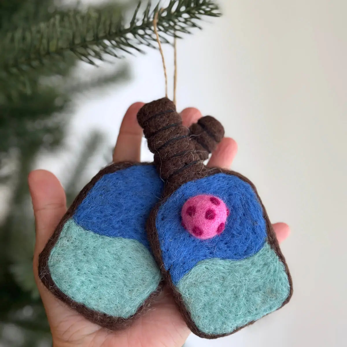 A hand holds two Felted Ornaments by Faire, shaped like lungs with blue and green sections and a pink polka-dotted circle. A tree branch appears in the background, highlighting these unique Christmas decorations.