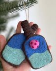 A hand holds two Felted Ornaments by Faire, shaped like lungs with blue and green sections and a pink polka-dotted circle. A tree branch appears in the background, highlighting these unique Christmas decorations.