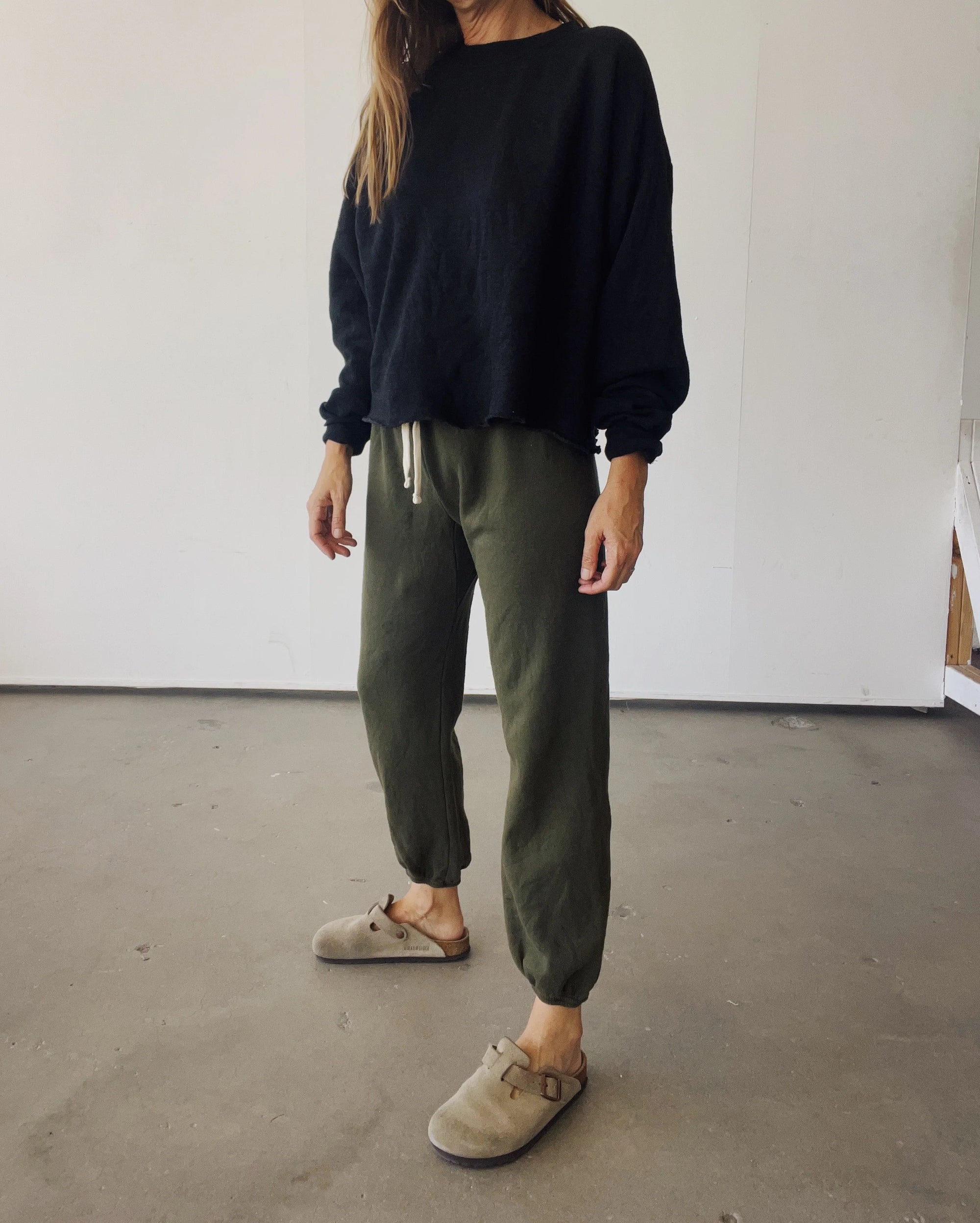 A person wearing a loose black long-sleeve shirt, Hey Gang&#39;s The Sweatpants in olive green, and taupe slip-on clogs stands indoors on a concrete floor. Their face is not visible.
