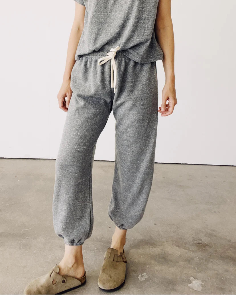 A person wearing The Sweatpants from Hey Gang is standing indoors. Their outfit includes a relaxed gray T-shirt and matching french terry sweatpants with an elastic waistband. With their hands at their sides, they also sport tan slip-on sandals against a plain white wall backdrop.