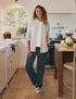 In a bright, modern kitchen, a woman wears Frank & Eileen’s Catherine Favorite Sweatpant with a white button-up shirt as she leans casually against a wooden island surrounded by white cabinetry, plants, and a bowl of fruit.