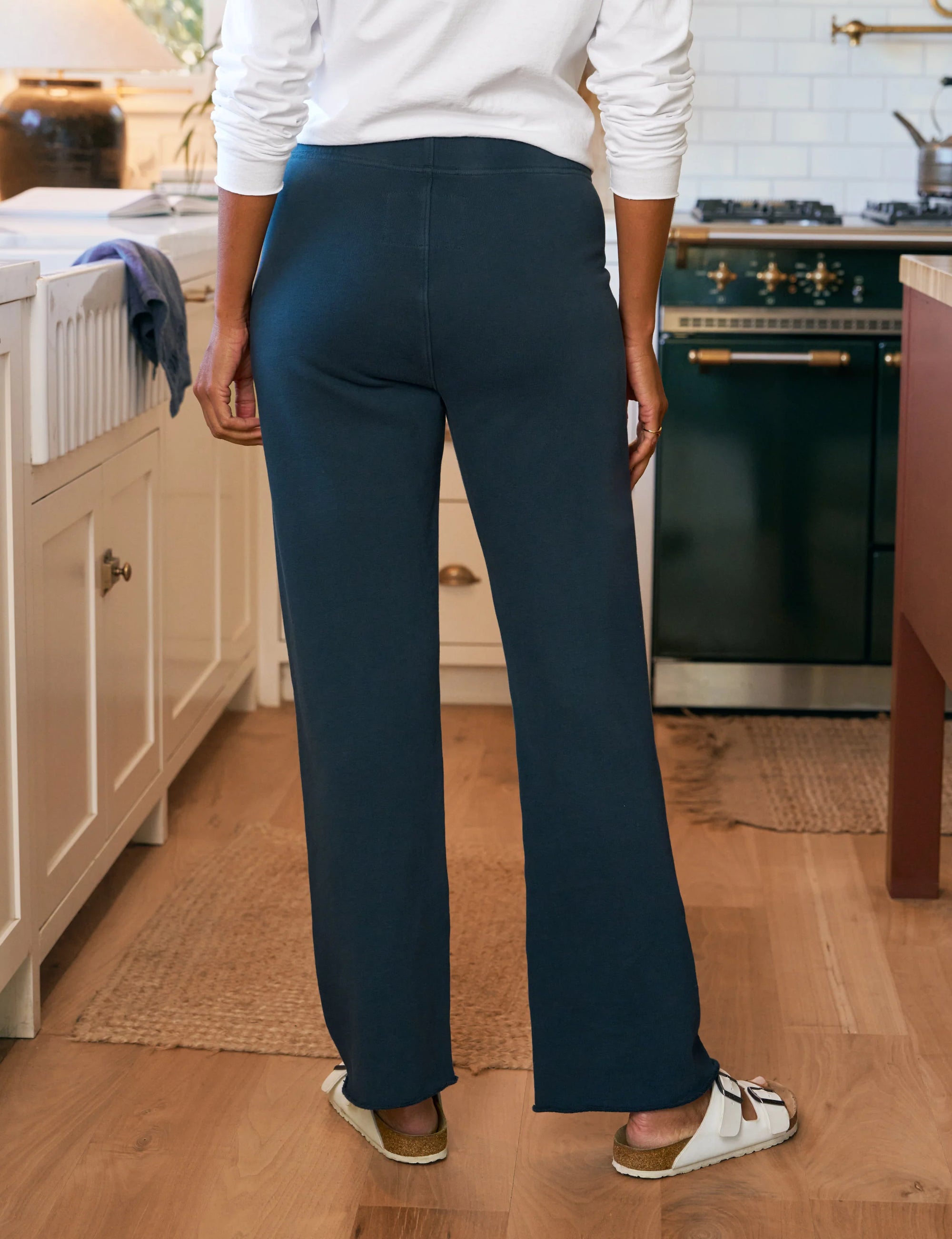 A person stands in a kitchen wearing a white long-sleeve shirt, white sandals, and Frank & Eileen Catherine Favorite Sweatpant in navy blue. The kitchen has light cabinets, a green stove, and wooden floors.