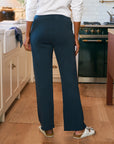 A person stands in a kitchen wearing a white long-sleeve shirt, white sandals, and Frank & Eileen Catherine Favorite Sweatpant in navy blue. The kitchen has light cabinets, a green stove, and wooden floors.