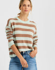 A woman with blonde hair tied back wears the Sloane Cashmere Boxy sweater by One Grey Day—a mint green and brown striped crewneck—styled with blue jeans, standing against a plain light background with one hand in her pocket.