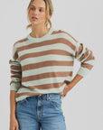 A person wears the Sloane Cashmere Boxy sweater by One Grey Day—featuring a loose knit, wide brown and light green stripes, and a crewneck—paired with blue jeans against a plain light gray background.