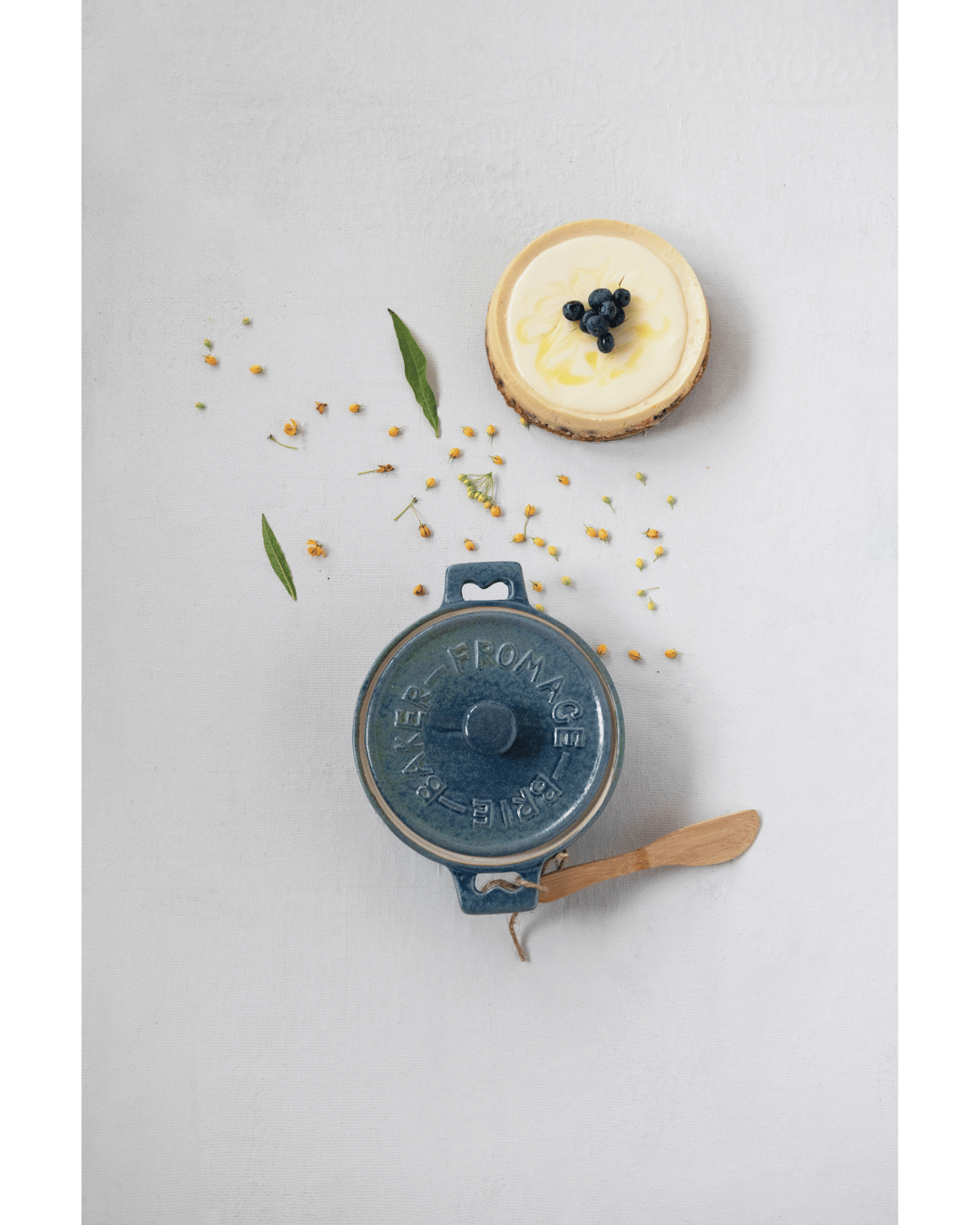 A Creative Co-op blue Stoneware Brie Baker with lid and bamboo spreader sits on a light surface. A lemon tart topped with blueberries appears at the top right, while small white flowers and green leaves are scattered around.