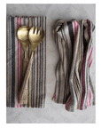 Two luxurious Woven Cotton Tea Towels from Creative Co-op in multi-color stripes elegantly complement any kitchen collection, with one towel folded beneath gold-toned salad servers and the other loosely gathered against a textured light gray background.