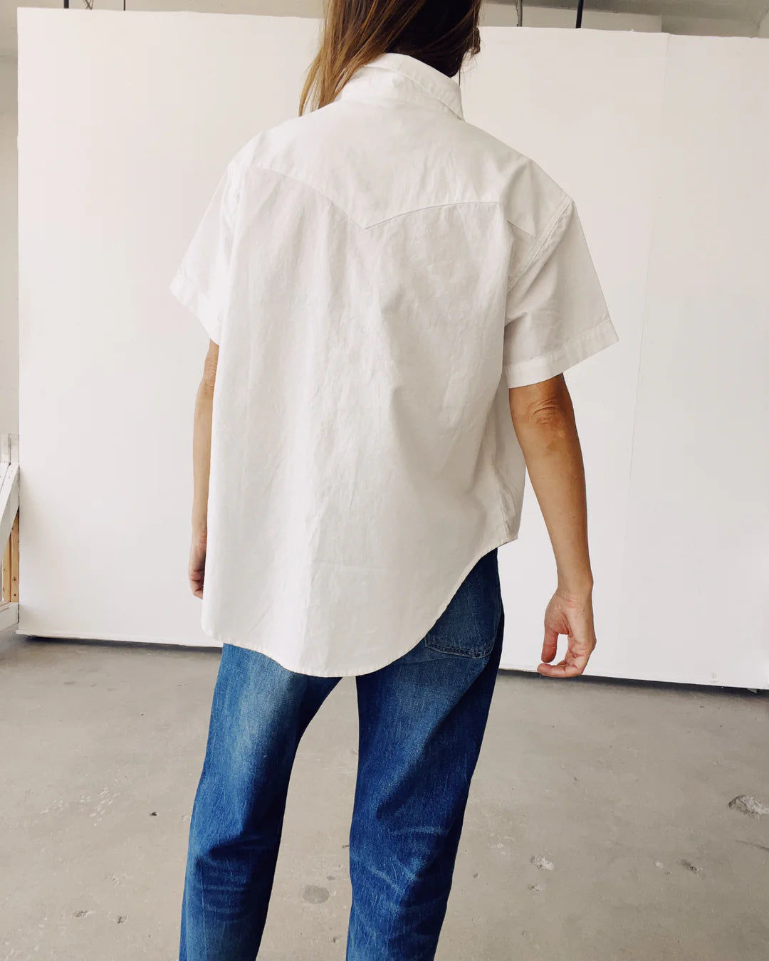 A person with long hair is facing away from the camera, wearing The Western Shirt by Hey Gang in white along with blue jeans. They are standing in a room with a light-colored wall and concrete floor.