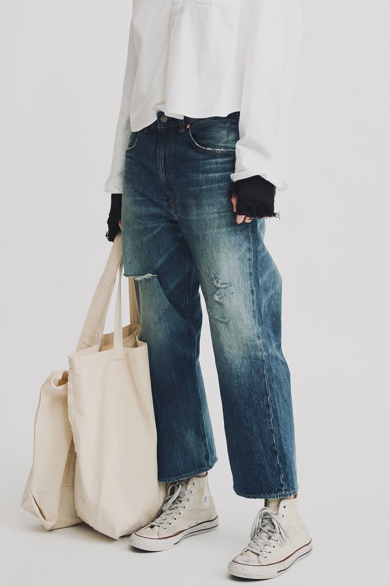 Model wears Denimist’s HAISLEY SLOUCH BF JEAN in blue, paired with a loose white long-sleeve shirt, white high-top sneakers, and carries a large beige tote bag against a plain white background.