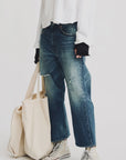 Model wears Denimist’s HAISLEY SLOUCH BF JEAN in blue, paired with a loose white long-sleeve shirt, white high-top sneakers, and carries a large beige tote bag against a plain white background.