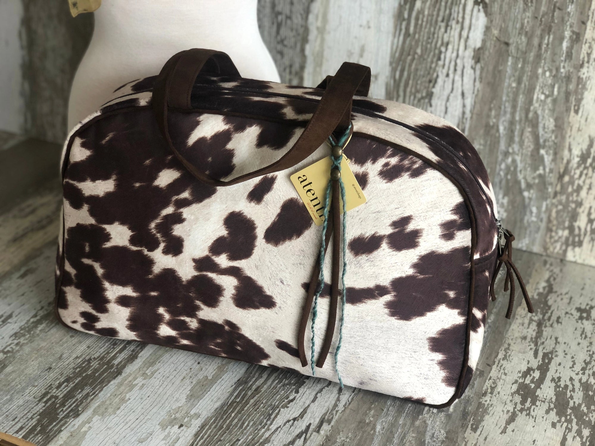 The Faire Oakley Overnighter - Faux Cowhide is a vegan rectangular weekender bag with a dark brown and white cowhide pattern, brown leather handles, and a yellow tag. Handmade in the USA, it's displayed on rustic wood with a weathered backdrop.