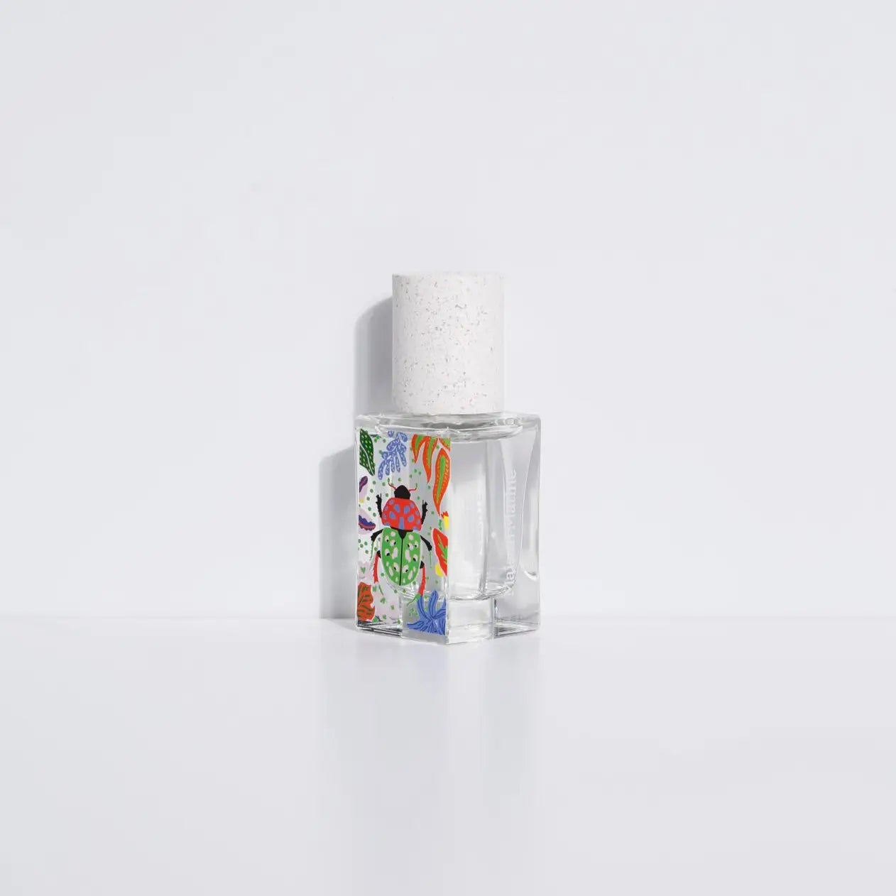 The Maison Matine Perfume by Faire features a clear bottle with a white speckled cap and vibrant abstract designs, displayed on a white surface against a plain background, suggesting bergamot notes in its delicate scent.