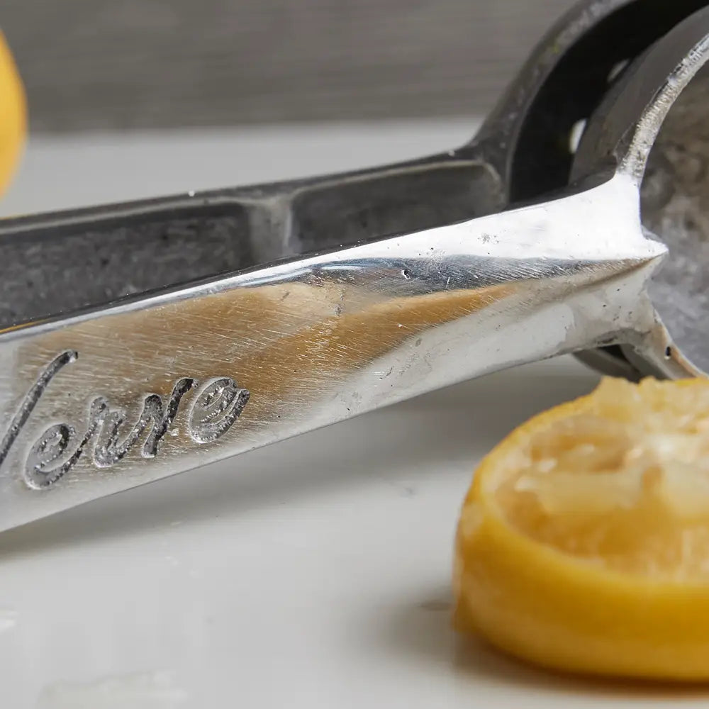 Close-up of Faire&#39;s Aluminum Hand Juicer with &quot;Verve&quot; engraved on the handle. A halved lemon, slightly crushed, sits on a white surface in the foreground. Ideal for extracting fresh lemon and lime juice, it&#39;s proudly made in Mexico. The background is softly blurred.