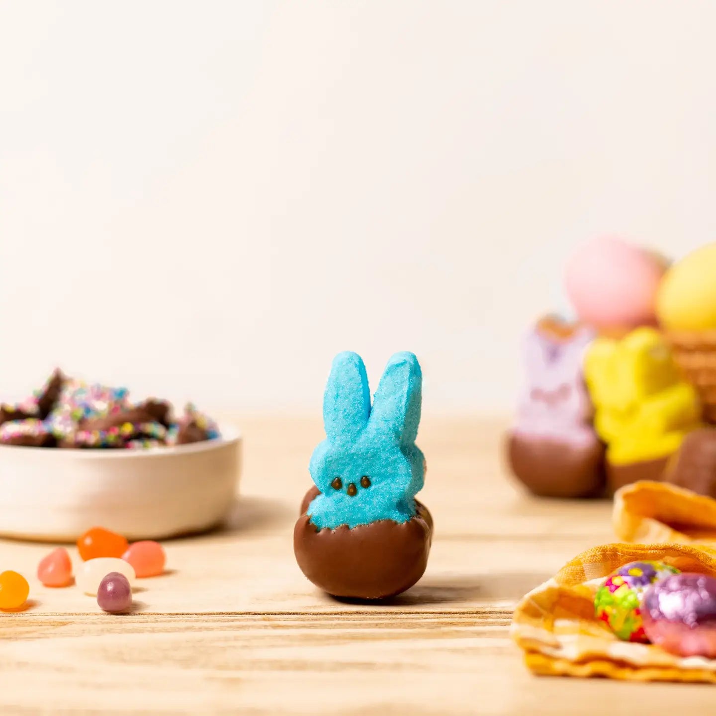 A Faire Easter Dipped Peeps marshmallow, coated in chocolate, rests on a wooden table with colorful candies, a bowl of sprinkles, and more marshmallow bunnies—an ideal treat for any Easter basket.
