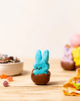 A Faire Easter Dipped Peeps marshmallow, coated in chocolate, rests on a wooden table with colorful candies, a bowl of sprinkles, and more marshmallow bunnies—an ideal treat for any Easter basket.