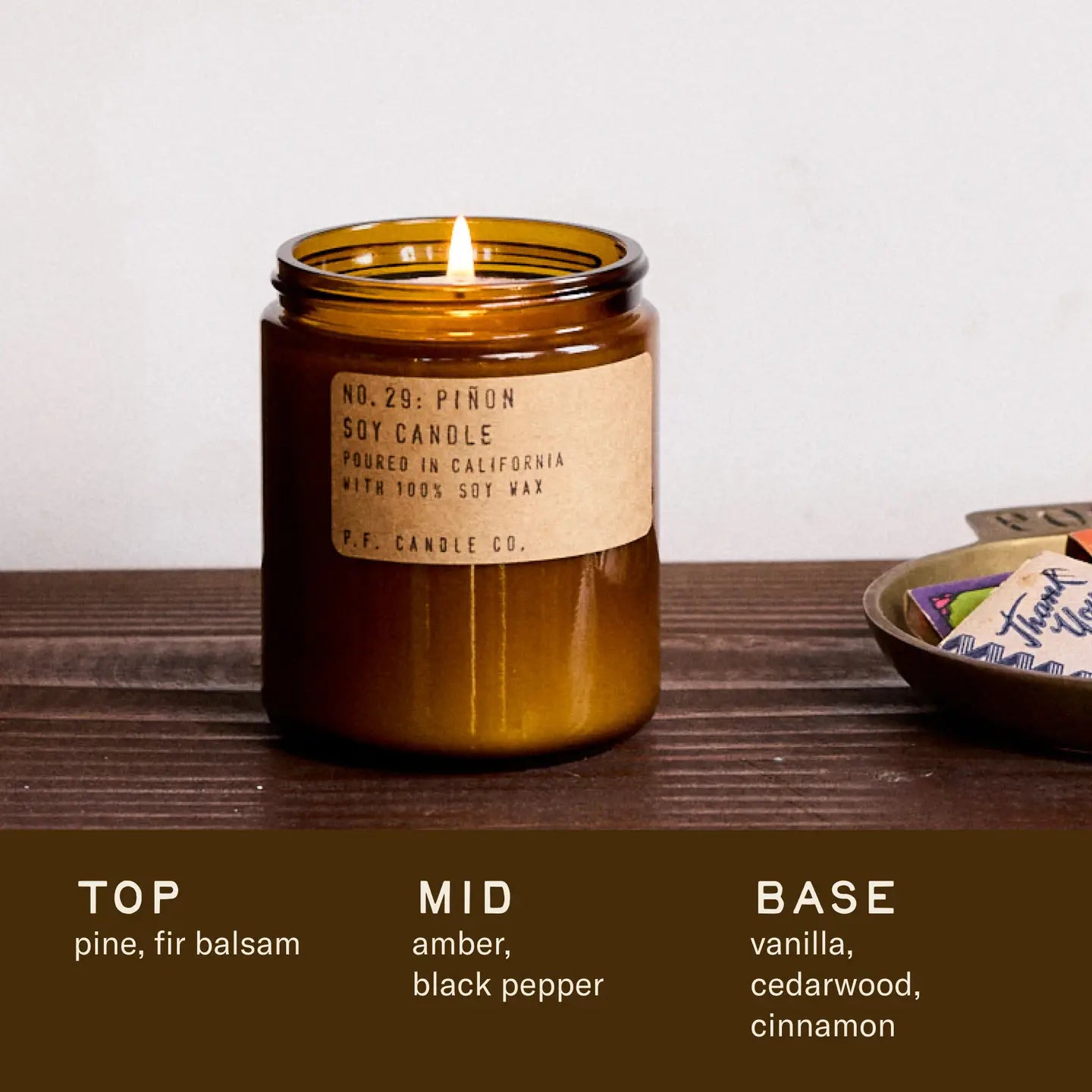A lit amber glass Faire PFC Candle rests on wood. Crafted with fine fragrance oils, it features scent notes of pine, fir balsam, amber, black pepper, vanilla, cedarwood, and cinnamon.