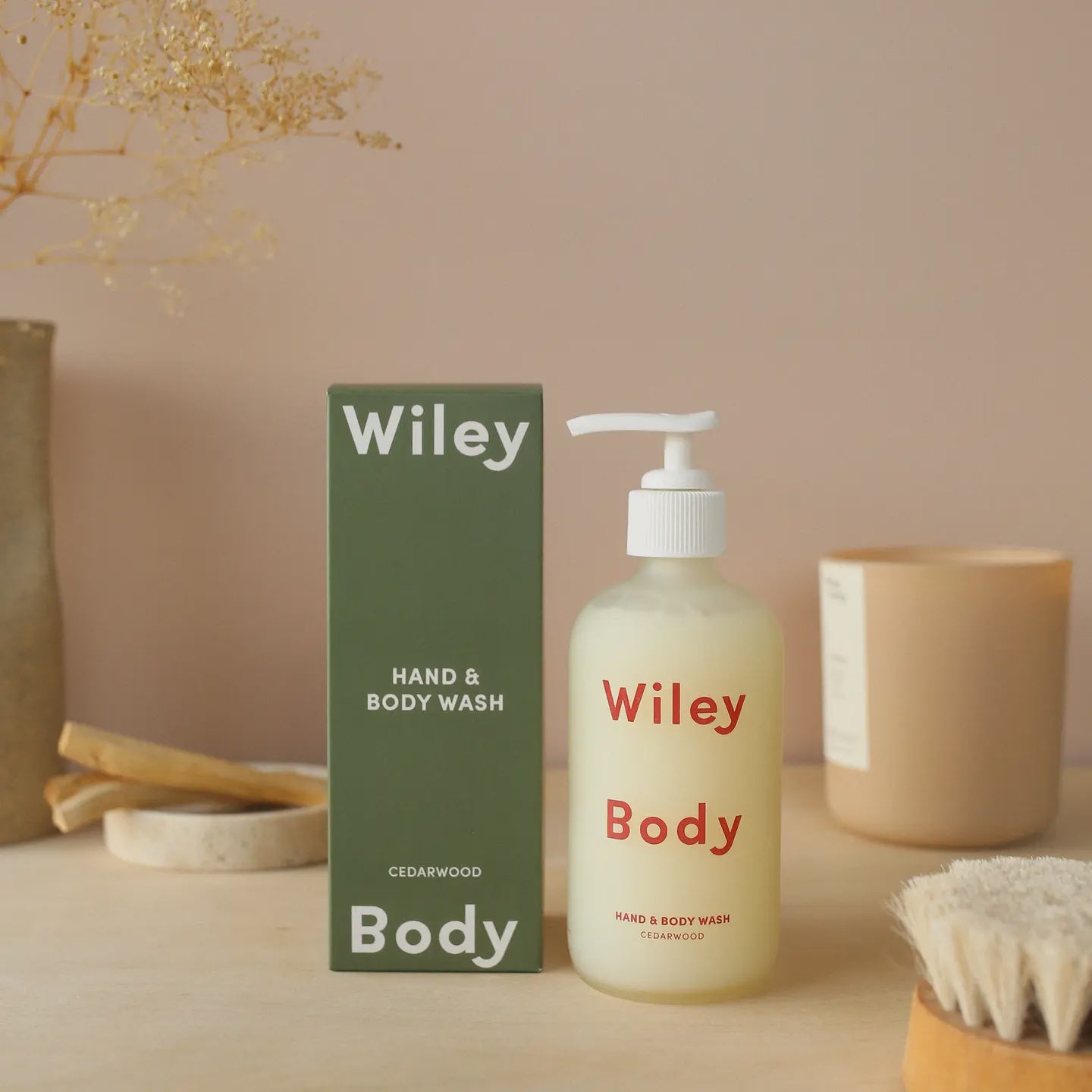 A frosted glass bottle of Faire's Wiley Hand & Body Wash with cedarwood scent is placed beside its green packaging, accompanied by a candle, brush, and decorative twigs on a light surface, creating an ambiance of natural serenity.