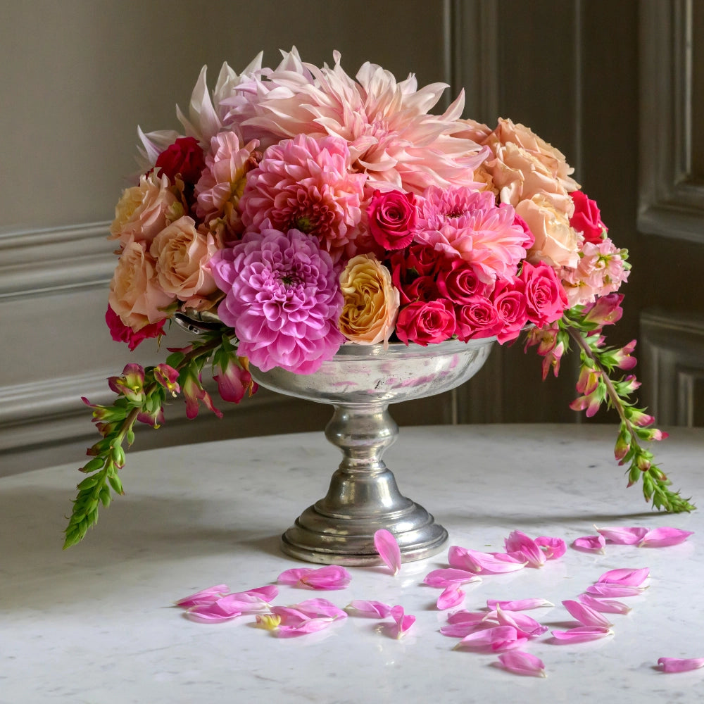 The Vintage 1795 Pedestal Bowl by Faire, handmade in Italy, overflows with pink, peach, and magenta blooms on a marble table with scattered pink petals completing the elegant display.
