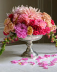 The Vintage 1795 Pedestal Bowl by Faire, handmade in Italy, overflows with pink, peach, and magenta blooms on a marble table with scattered pink petals completing the elegant display.
