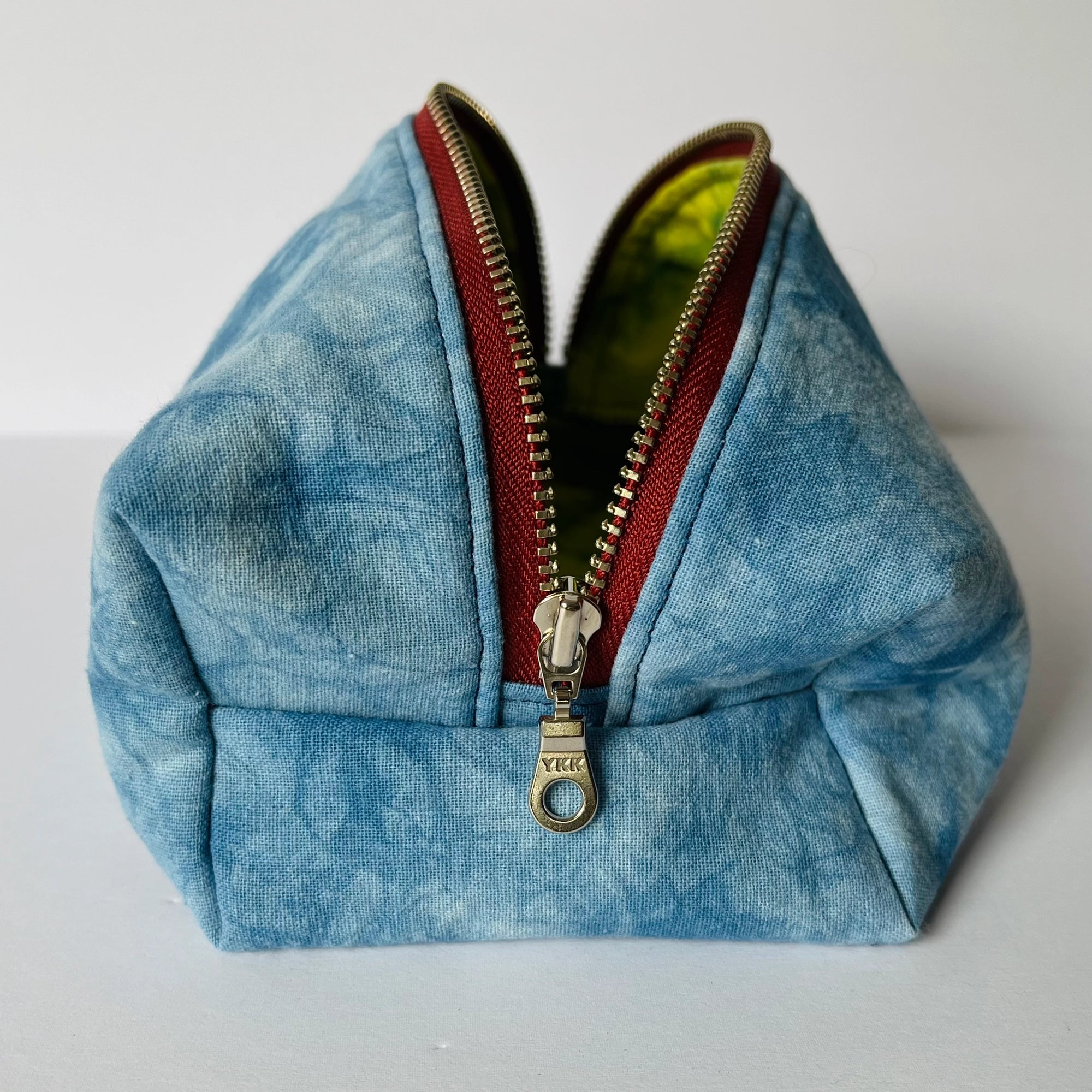 The Faire Dyed Toiletries Bag is a small, blue hand-dyed cotton pouch with a marbled pattern, red-trimmed metal zipper, and yellow-green lining—ideal for organizing your desk or daily essentials. Shown upright on a white surface.