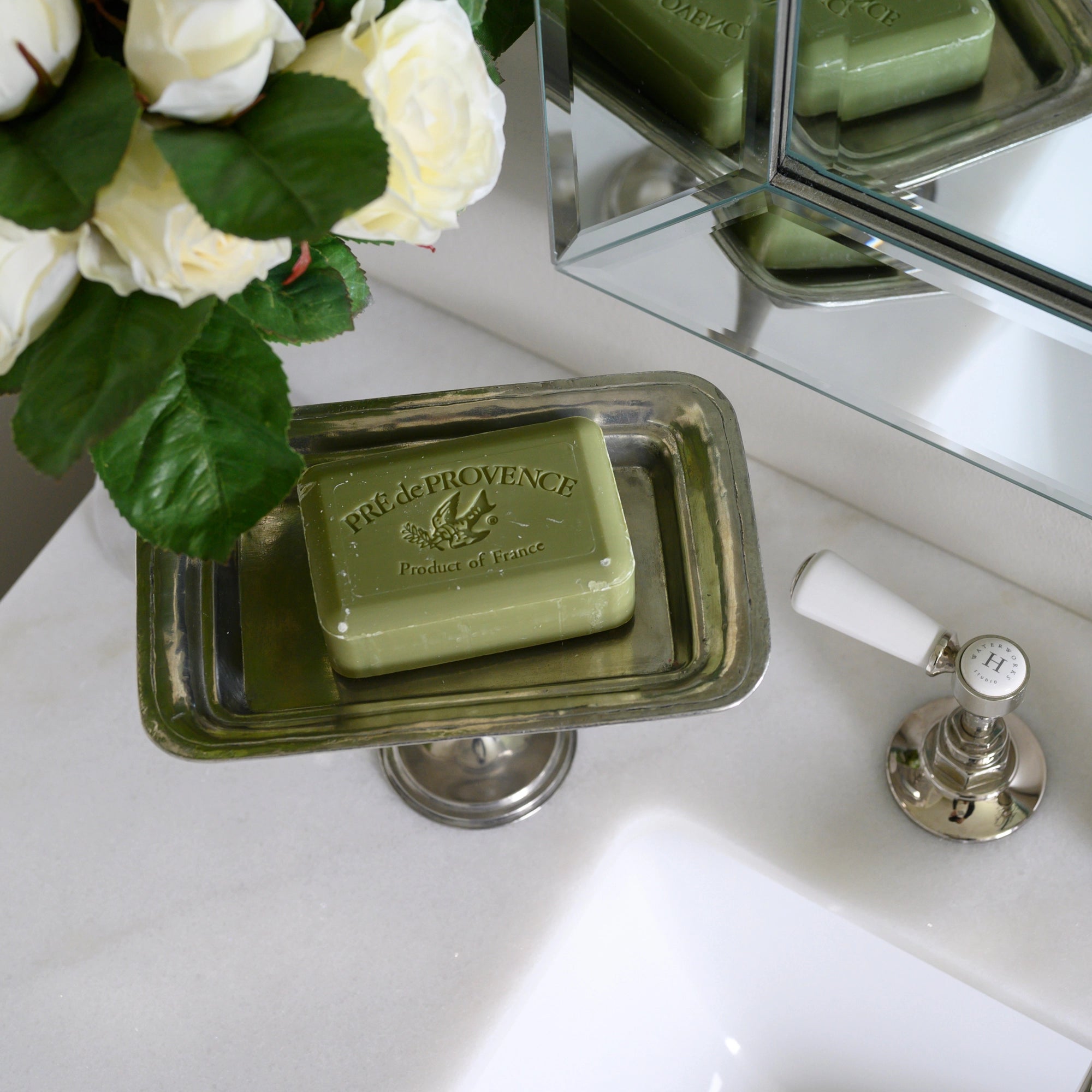 A green soap bar sits on a Faire Vintage Rec Pedestal Dish beside a white sink, accented by white roses and a chrome faucet, while a mirror reflects the elegant scene.