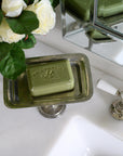 A green soap bar sits on a Faire Vintage Rec Pedestal Dish beside a white sink, accented by white roses and a chrome faucet, while a mirror reflects the elegant scene.