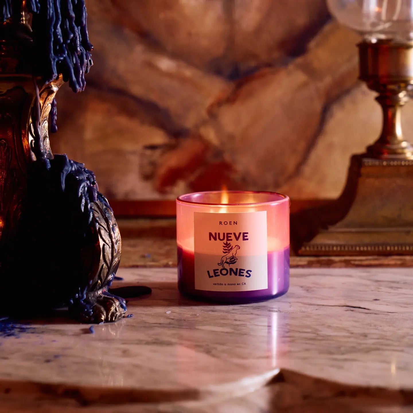 A lit purple &quot;Roen Candles&quot; by Faire sits on marble, made with a coconut-soy wax blend. It&#39;s in a decorative holder with dripped blue wax, next to a vintage lamp against an art nouveau mansion&#39;s warm-toned, textured wall.