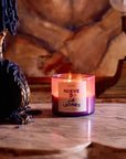 A lit purple "Roen Candles" by Faire sits on marble, made with a coconut-soy wax blend. It's in a decorative holder with dripped blue wax, next to a vintage lamp against an art nouveau mansion's warm-toned, textured wall.