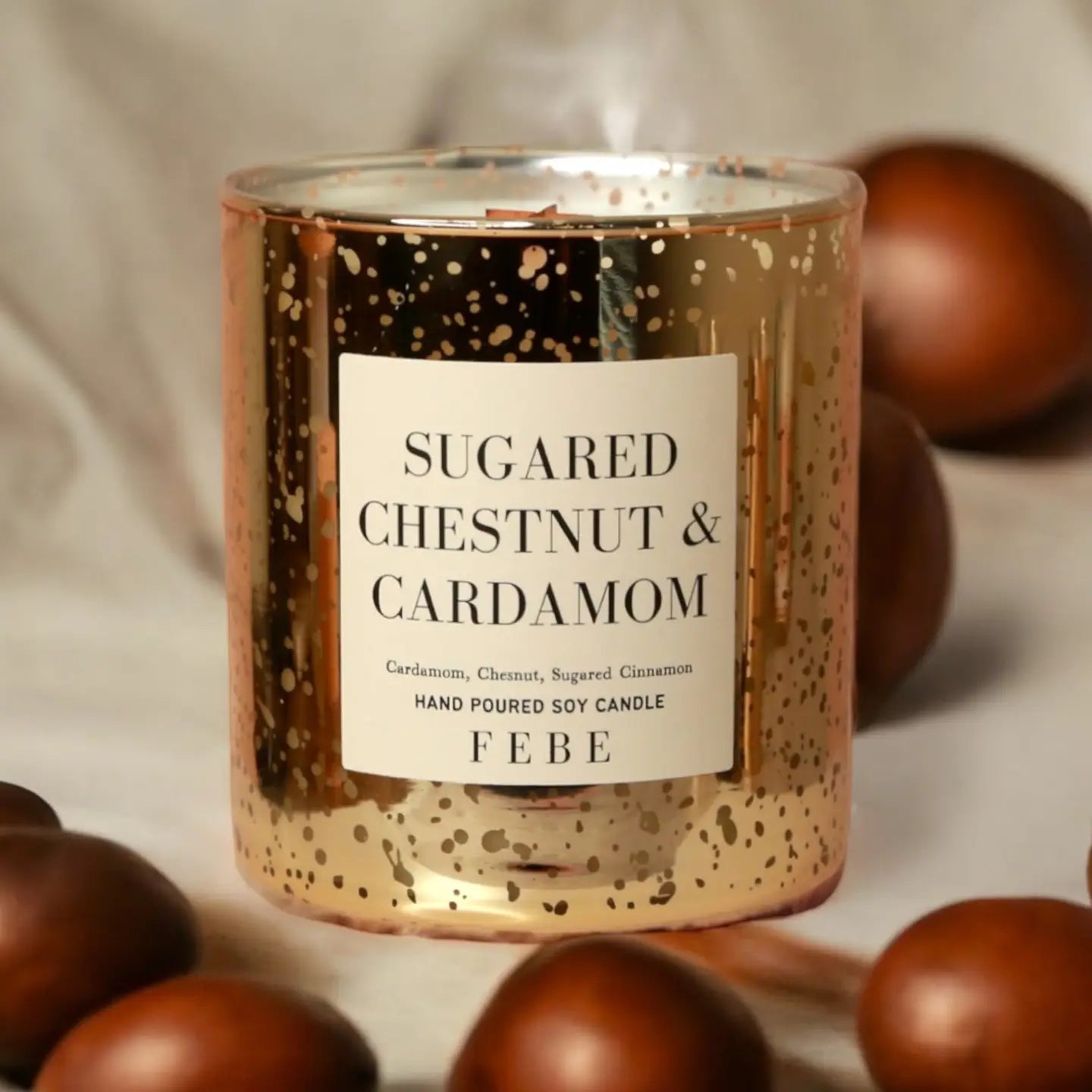 A gold-speckled glass candle labeled "Sugared Chestnut & Cardamom" from FEBE Candles by Faire shines warmly, surrounded by scattered brown chestnuts and the comforting essence of an Apple Cider Donut on a soft, beige background.