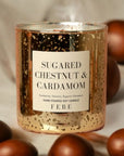 A gold-speckled glass candle labeled "Sugared Chestnut & Cardamom" from FEBE Candles by Faire shines warmly, surrounded by scattered brown chestnuts and the comforting essence of an Apple Cider Donut on a soft, beige background.