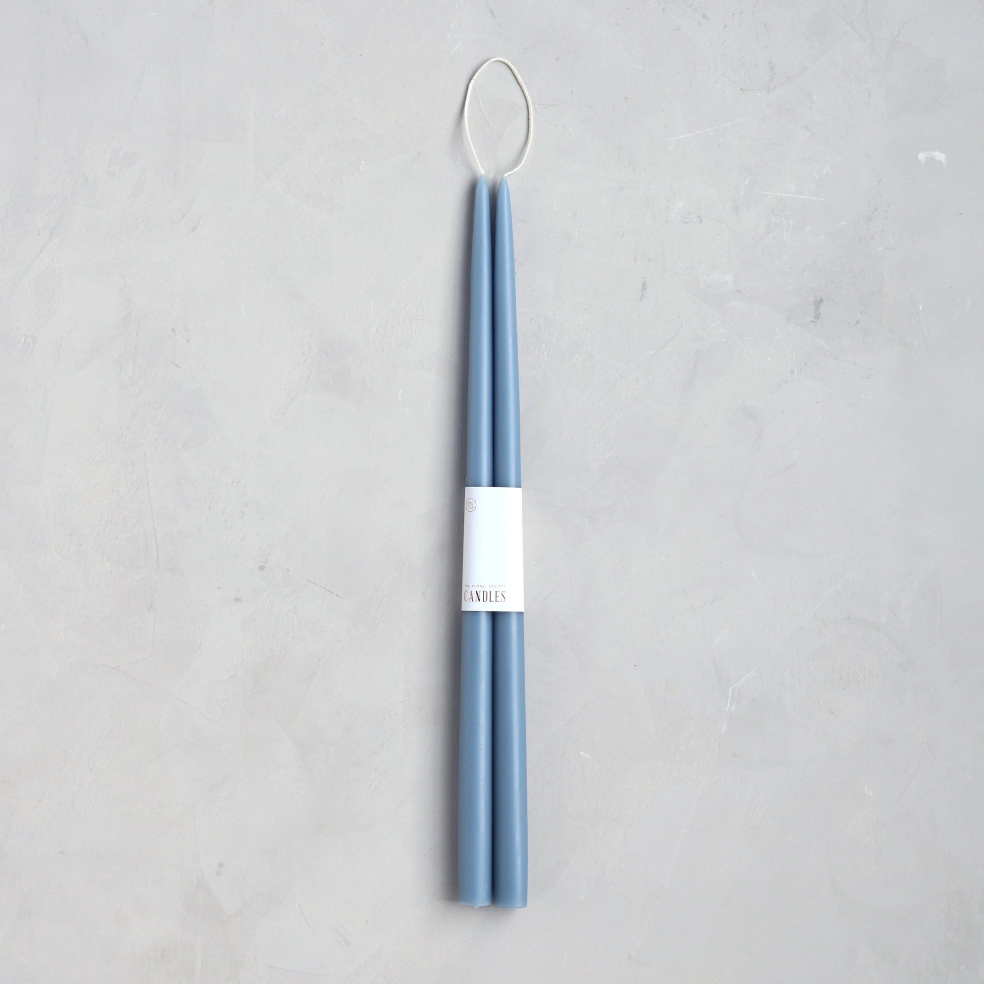 The Floral Society 18" Taper Candle Pair in blue features tall, slender candles bound with a white band and topped with a looped wick—an elegant touch for any home décor. Set on a light gray textured surface.