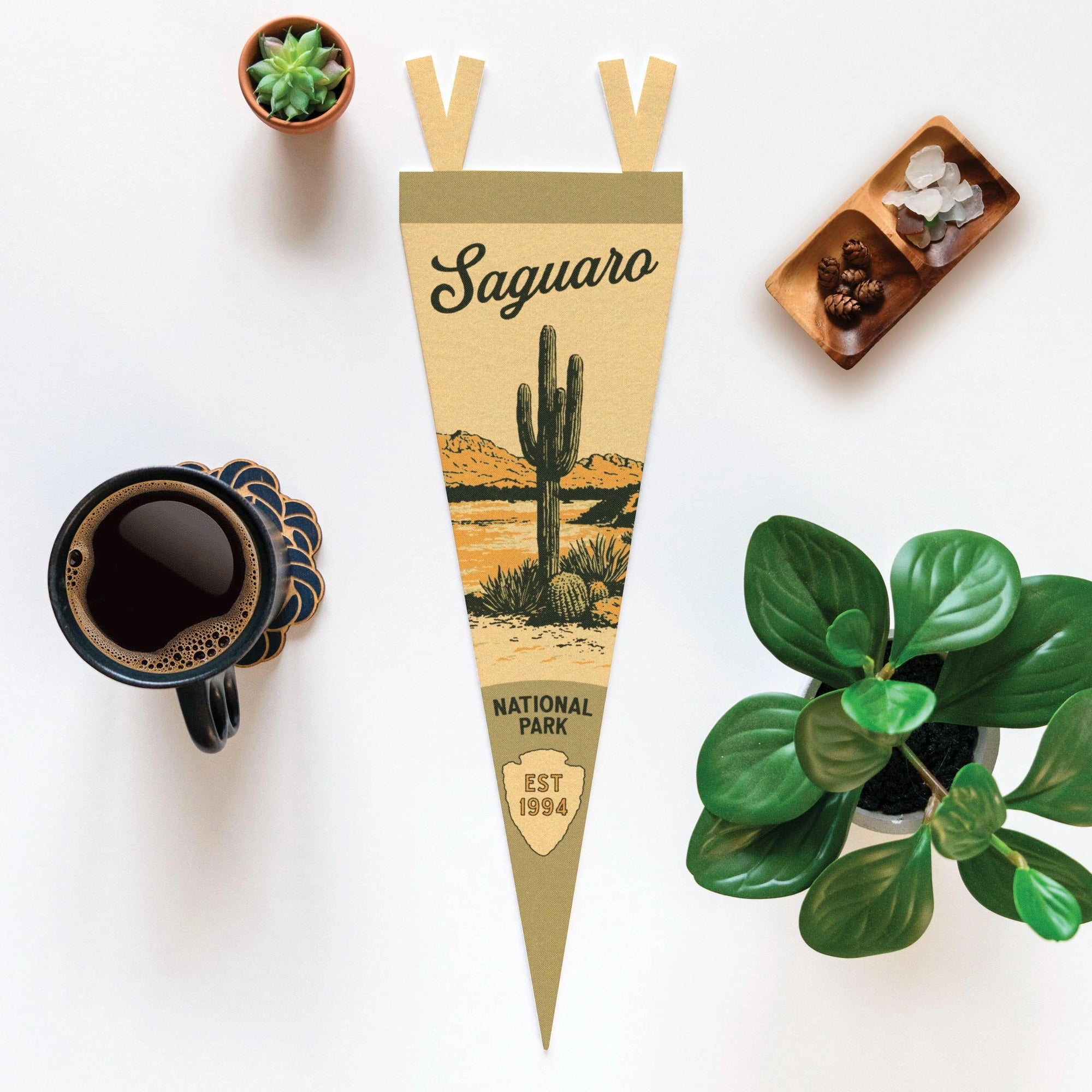 The Faire Saguaro Ntnl Park Pennant is shown on a white surface beside coffee, a succulent, a leafy plant, and a tray of crystals.