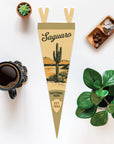 The Faire Saguaro Ntnl Park Pennant is shown on a white surface beside coffee, a succulent, a leafy plant, and a tray of crystals.