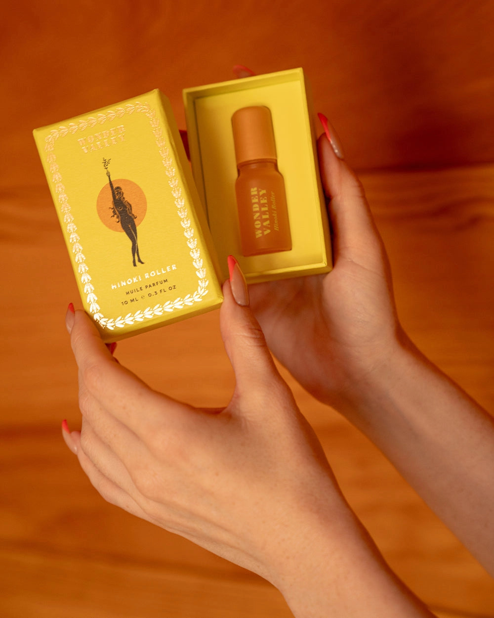 A person with manicured nails holds a yellow box containing Faire’s Hinoki Roll On—a small essential oil roller with a refreshing hinoki cedar scent—against a wooden background.