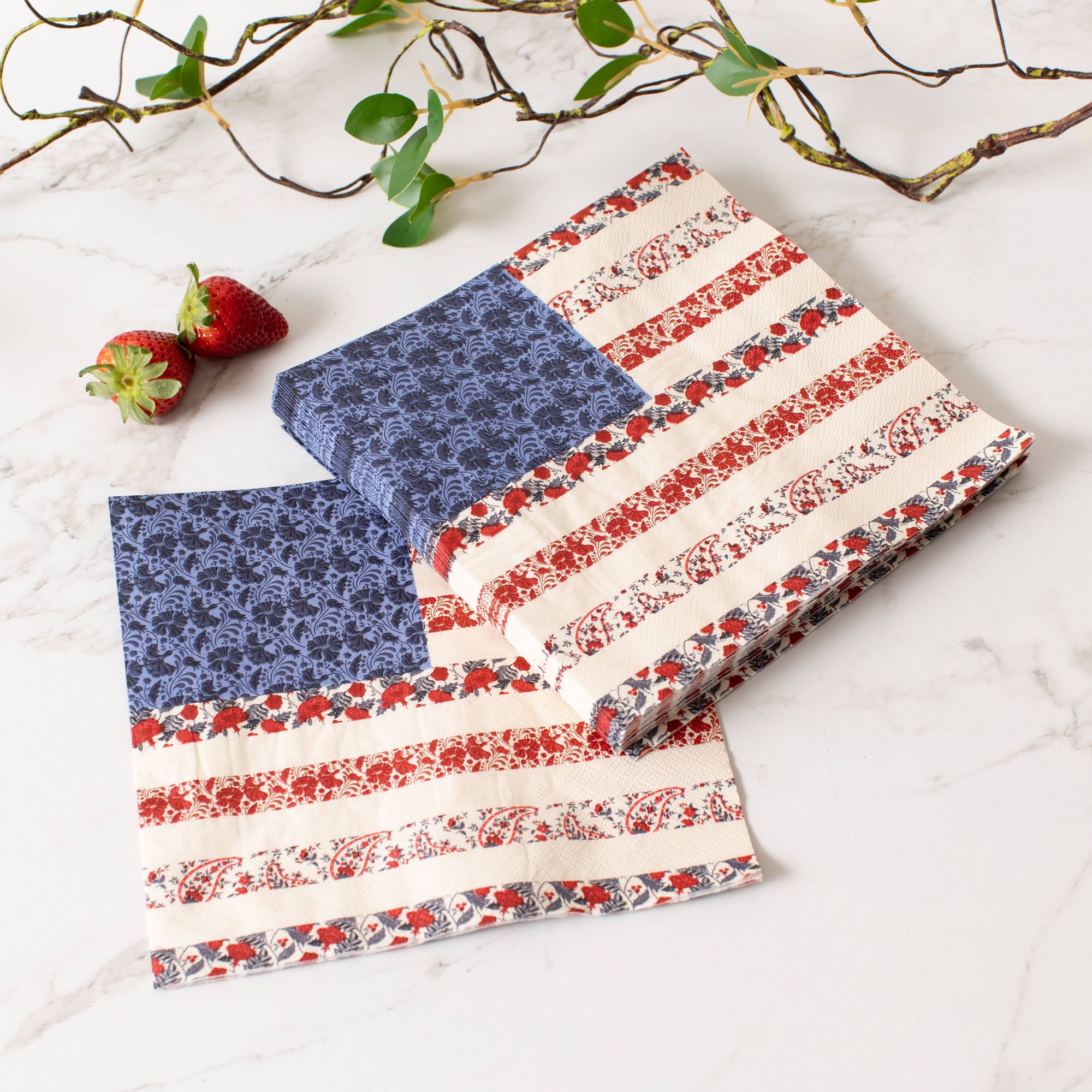 Two Faire Americana Flag Paper Napkins, featuring blue floral and red-striped accents inspired by the American flag, are displayed folded on a marble surface beside two strawberries and a leafy branch.