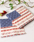 Two Faire Americana Flag Paper Napkins, featuring blue floral and red-striped accents inspired by the American flag, are displayed folded on a marble surface beside two strawberries and a leafy branch.