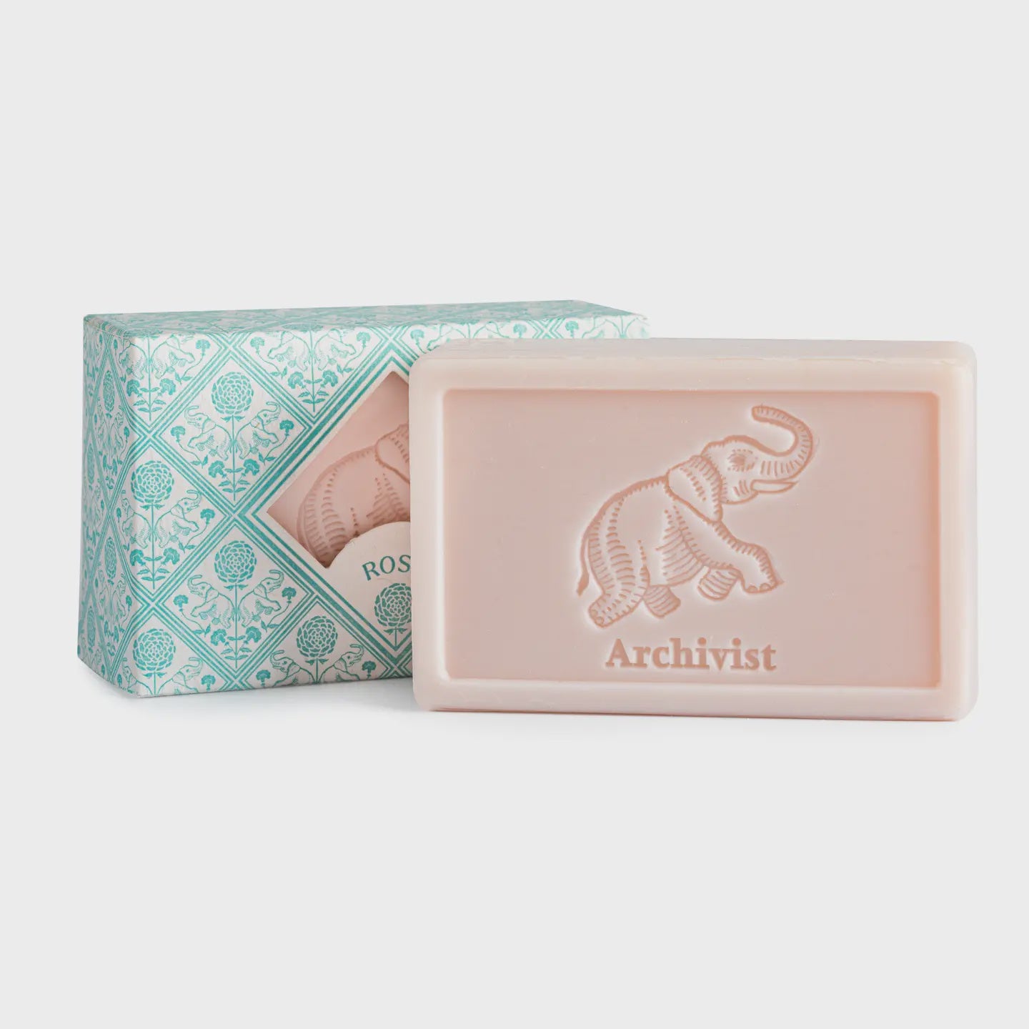 A rectangular pale pink soap bar by Faire with a letterpressed elephant and "Savon d'Archivist" embossed on it, displayed in front of a decorative teal and white "Rose" box, captures the charm of Provence.