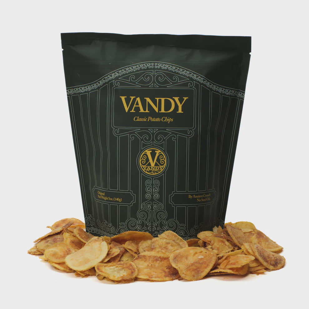 A bag of Vandy Chips from Faire is displayed, showcasing a dark green design with gold lettering. The handmade bag is surrounded by a pile of crispy potato chips that are free from pesticides, set against a white background.