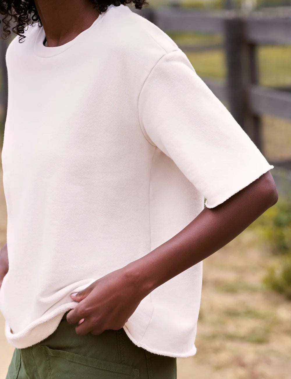 Woman in a casual crew neck tee by Frank &amp; Eileen with green pants stands outdoors near a rustic wooden fence.