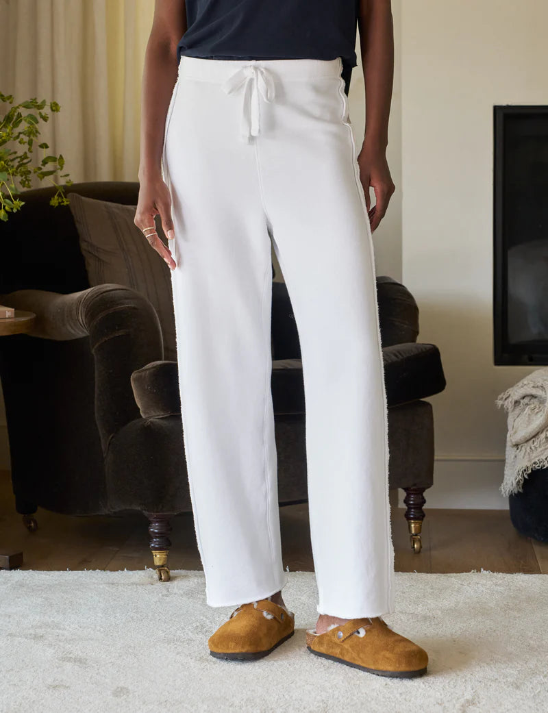 A person wearing cozy white Bella Sweatpants by Frank & Eileen and brown slippers stands on a light carpet. An upholstered brown armchair, a plant, and a fireplace add to the room's cozy ambiance.