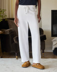 A person wearing cozy white Bella Sweatpants by Frank & Eileen and brown slippers stands on a light carpet. An upholstered brown armchair, a plant, and a fireplace add to the room's cozy ambiance.