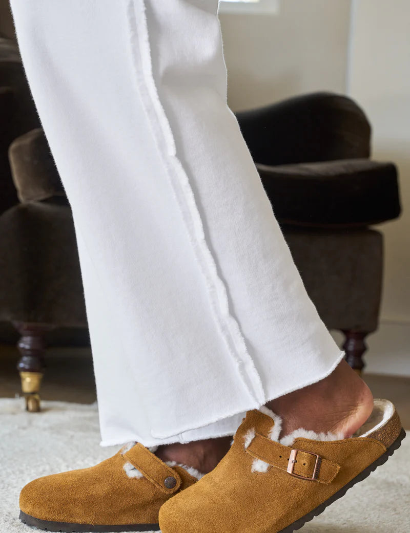 A person in Bella Sweatpants by Frank &amp; Eileen and brown shearling-lined clogs stands on a light-colored carpet, with a dark cushioned chair in the background, creating a cozy vibe.