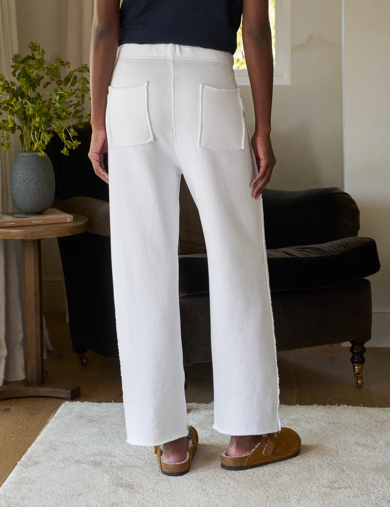 Rear view of a person in Frank &amp; Eileen&#39;s Bella Sweatpants with brown shoes, standing on a light rug. A dark sofa and side table with a vase of greenery complete the cozy background setting.